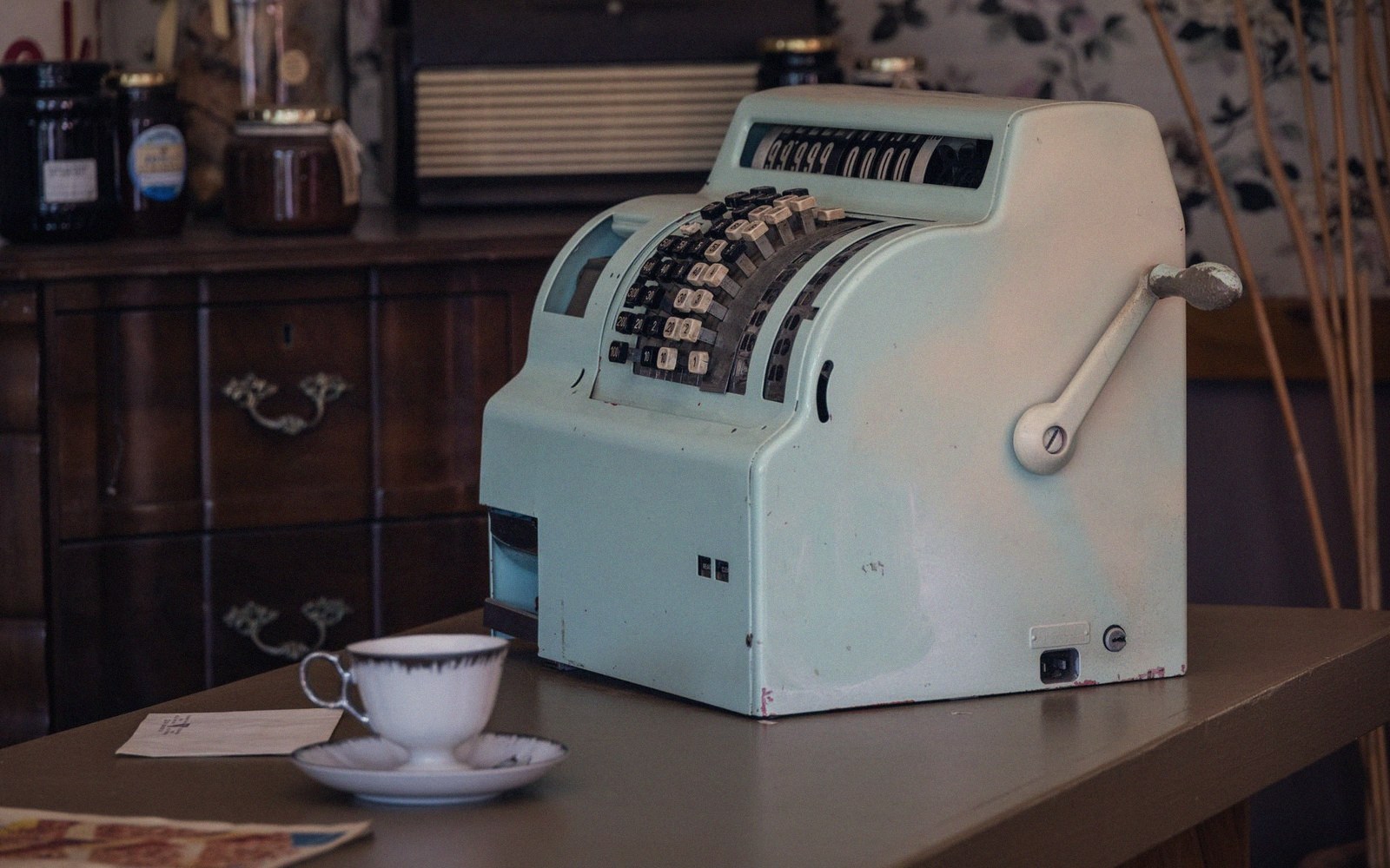 Vintage mint-green mechanical cash register with hand crank sits beside a teacup